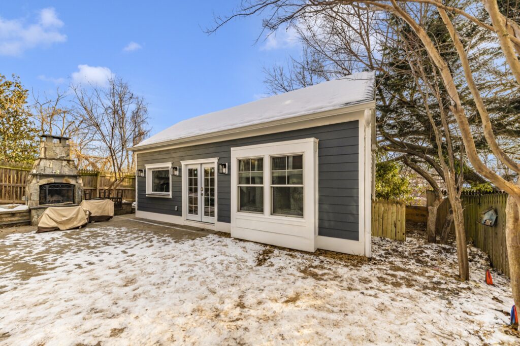 A small modern gray Tiny Home with white trim and large windows sits in a backyard dusted with snow, surrounded by bare trees and a wooden fence. An outdoor fireplace and grill are visible on the left.